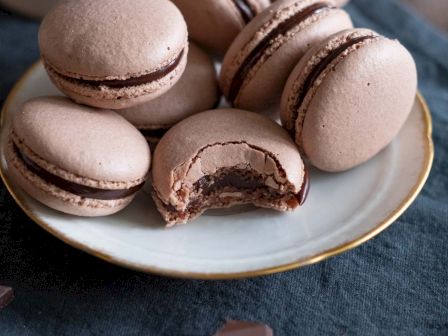 Delicate pink macarons with chocolate filling on a plate, a bite reveals rich ganache; chocolate shards and coffee cup in the background, yum.