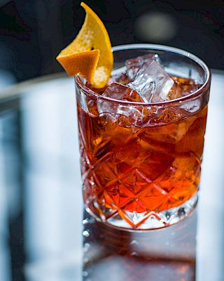 A crystal-cut glass with a dark amber cocktail and a orange wedge garnish on a reflective surface; elegant, classic bar scene.