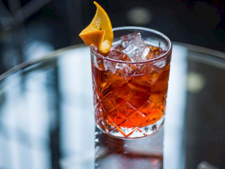 A crystal-cut glass with a dark amber cocktail and a orange wedge garnish on a reflective surface; elegant, classic bar scene.