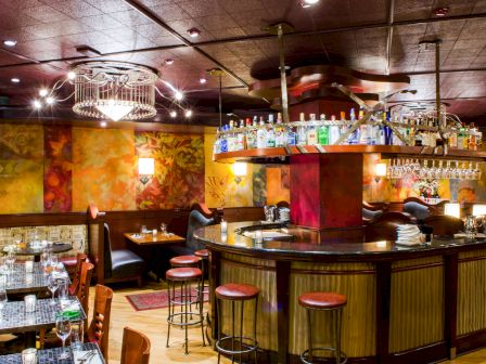A cozy bar with a circular wooden counter, hanging glass shelves, colorful walls, and intimate tables for diners, warm lighting, and stools.