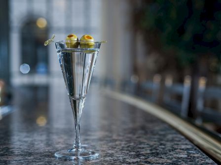 Two elegant martini glasses on a speckled bar, each topped with olive skewers, set against a blurred, ambient bar backdrop. Top it at 140 characters, always ending the sentence.