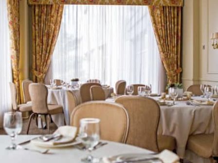 Elegant dining room with round tables, white tablecloths, gold chairs, chandeliers, and heavy floral drapes framing a bright window.