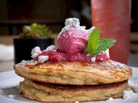 A stack of golden pancakes topped with pink ice cream, mint leaf, and crushed toppings on a white plate, with a red drink in the background.
