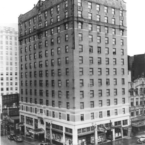 A black-and-white photo of a tall, early 20th-century brick building on a busy city street, with storefronts on the ground floor and cars nearby.