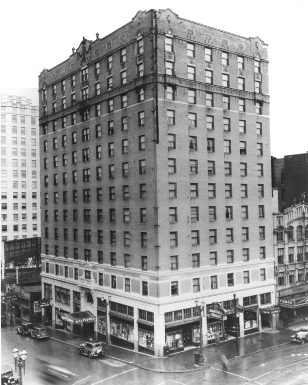 A black-and-white photo of a tall, early 20th-century brick building on a busy city street, with storefronts on the ground floor and cars nearby.