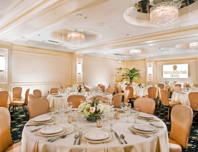 A formal banquet hall with round tables set for a meal, elegant white tablecloths, centerpieces, and chandeliers, ready for a reception.