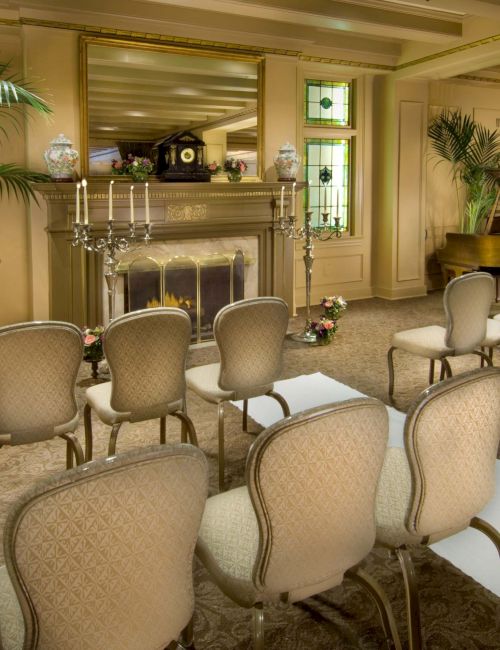 A formal chapel setup with beige upholstery chairs, a grand piano, a lit fireplace, candelabras, green plants, and an aisle leading to a small altar area.