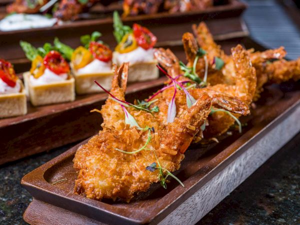 Golden fried shrimp skewers on a wooden tray, garnished with greens, plus bite-sized appetizers with cream and toppings in the background.