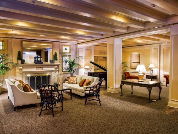 A spacious hotel lobby with warm lighting, a fireplace, comfortable seating, potted plants, a grand piano, and a few tables and lamps, inviting and elegant.
