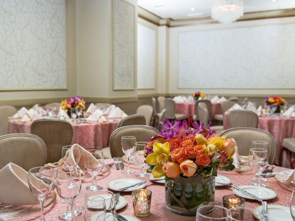 A banquet hall set for a formal event with round tables, pink tablecloths, floral centerpieces, and neatly arranged place settings.
