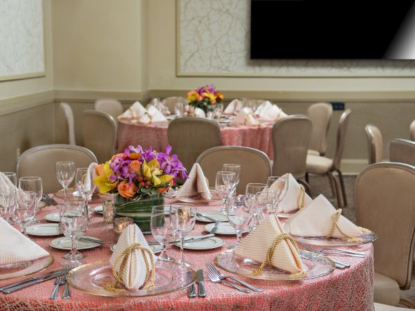 A banquet hall set for a formal dinner with round tables, pink tablecloths, folded napkins, glassware, and colorful flower centerpieces.