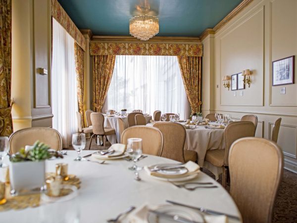 A refined dining room with round tables, cream chairs, elegant place settings, soft lighting, and floral drapes by tall windows.