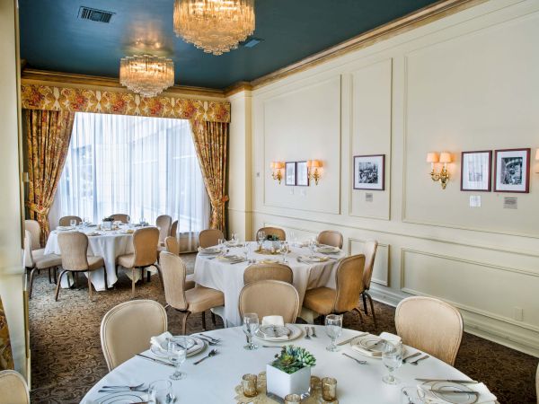 A elegant dining room with white paneled walls, chandeliers, round tables set with white linens, and beige upholstered chairs.