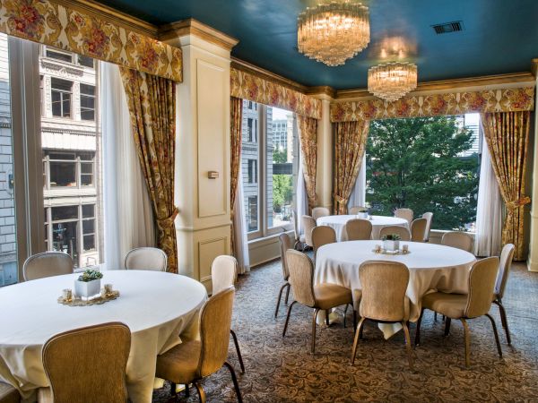Elegant dining room with round tables, cream chairs, chandeliers, and large windows; gold drapes and a teal ceiling create a luxurious, airy space.