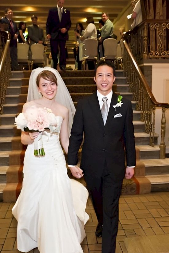 A newlywed couple in wedding attire walk hand-in-hand down a staircase in a hotel lobby, smiling amid guests and plants.