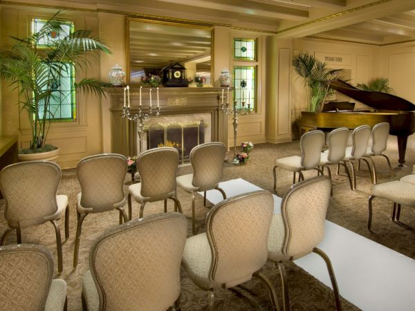 A formal ceremony setup with rows of beige chairs facing a mantle altar, candlelit, potted plants, a grand piano, and soft warm lighting.