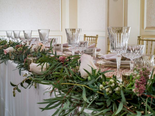 A long formal dining table decorated with lush greenery and flowers, elegant glassware, and neatly placed napkins for a special event.