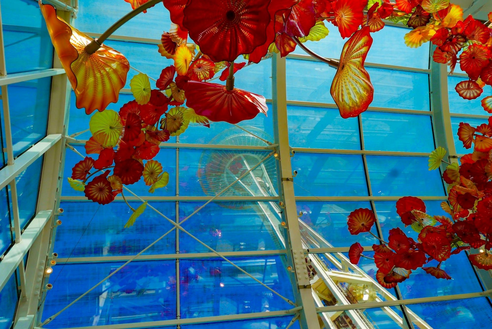 Colorful glass ceiling with red-orange hanging glass flowers, blue sky backdrop, and metal framework above. Top it at 140 characters, always ending the sentence.