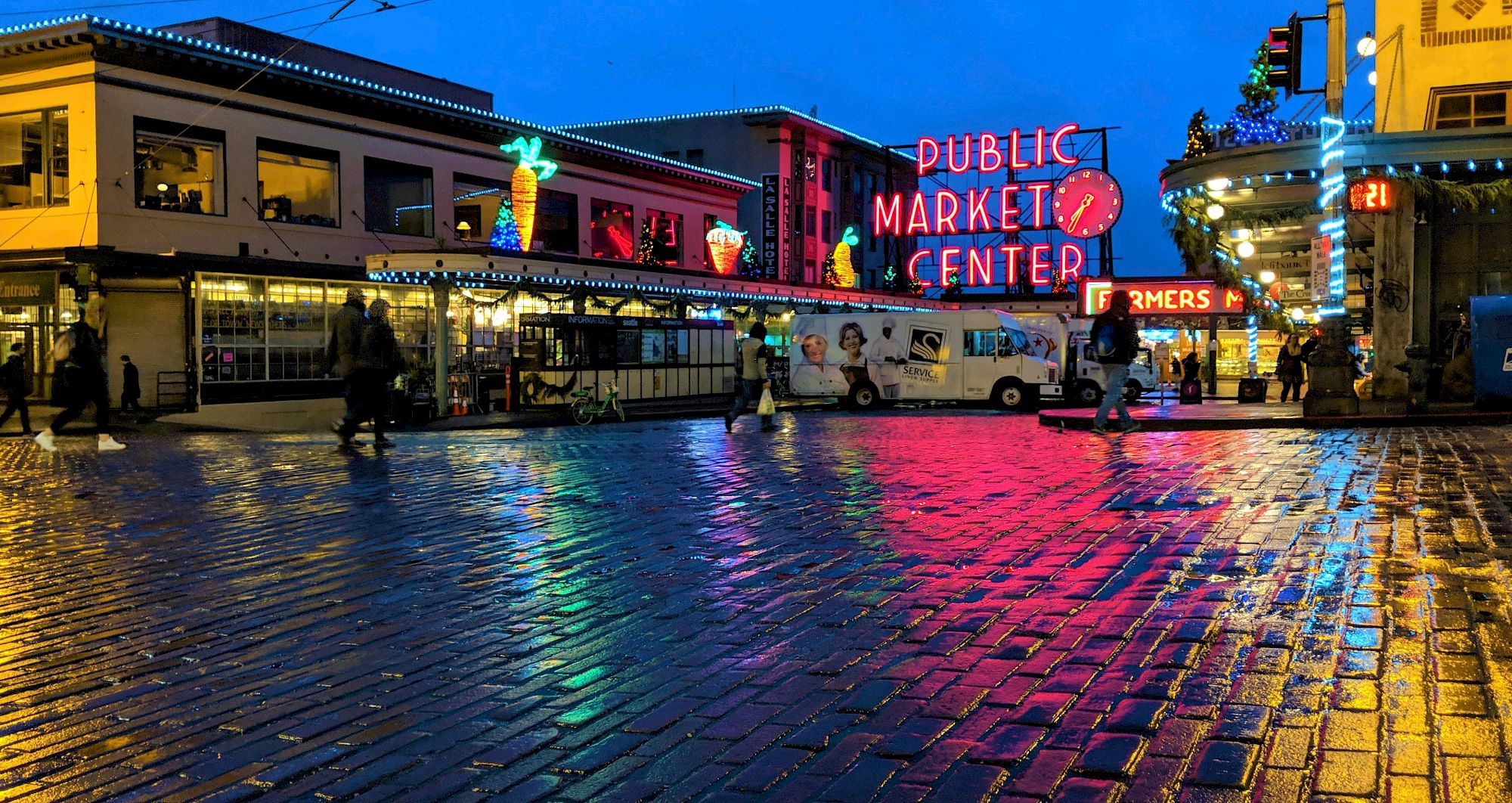A lively night scene at Public Market Center, neon signs glow, colorful reflections on wet cobblestones, vendors and crowds beneath bright storefronts.