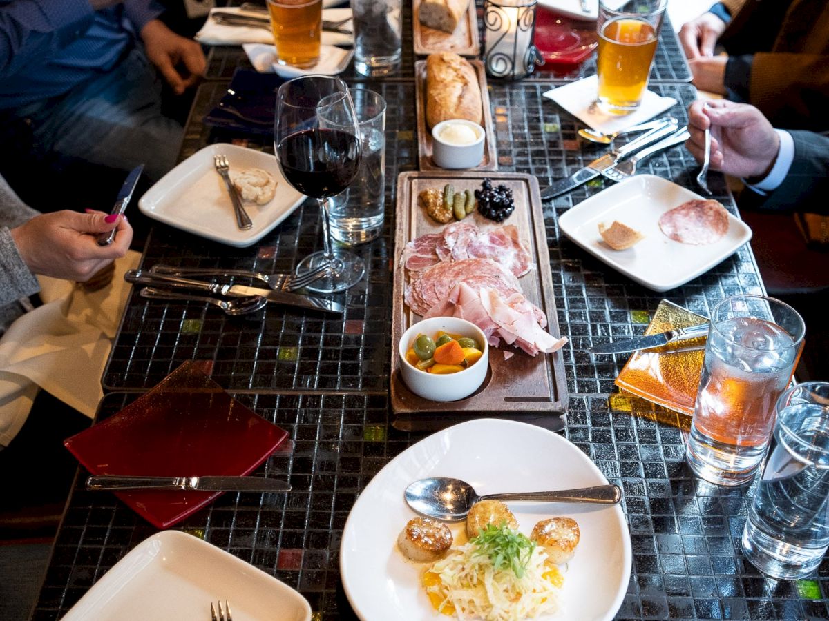 A long dinner table with assorted plates, drinks, and several people sharing a meal; various appetizers and a central dish at center.