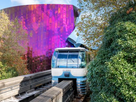 A light blue funicular or tram sits on tracks beside a lush green hedge, with a striking purple-pink glassy building in the background.
