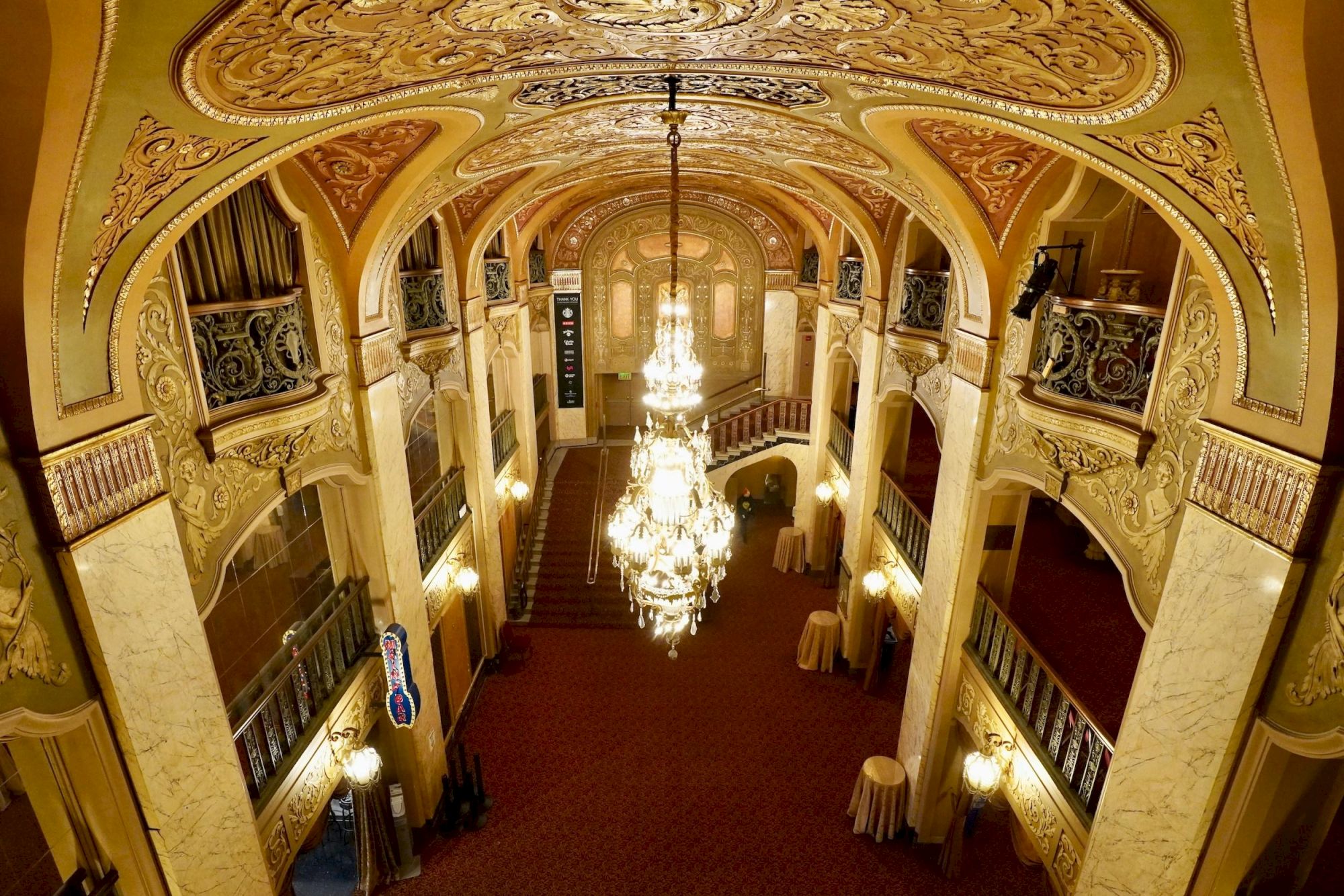An ornate theater lobby with gilded arches, a grand chandelier, balconies, and warm lighting, viewed from above as guests arrive.