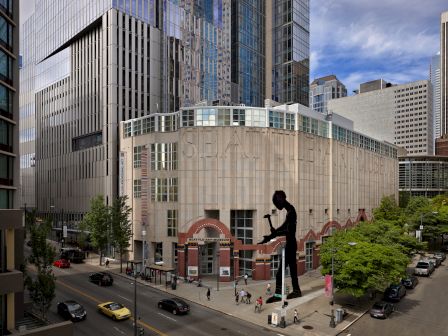 A busy city street with tall modern buildings, a large white circular building, and a silhouette statue of a person on a pedestal by the corner.