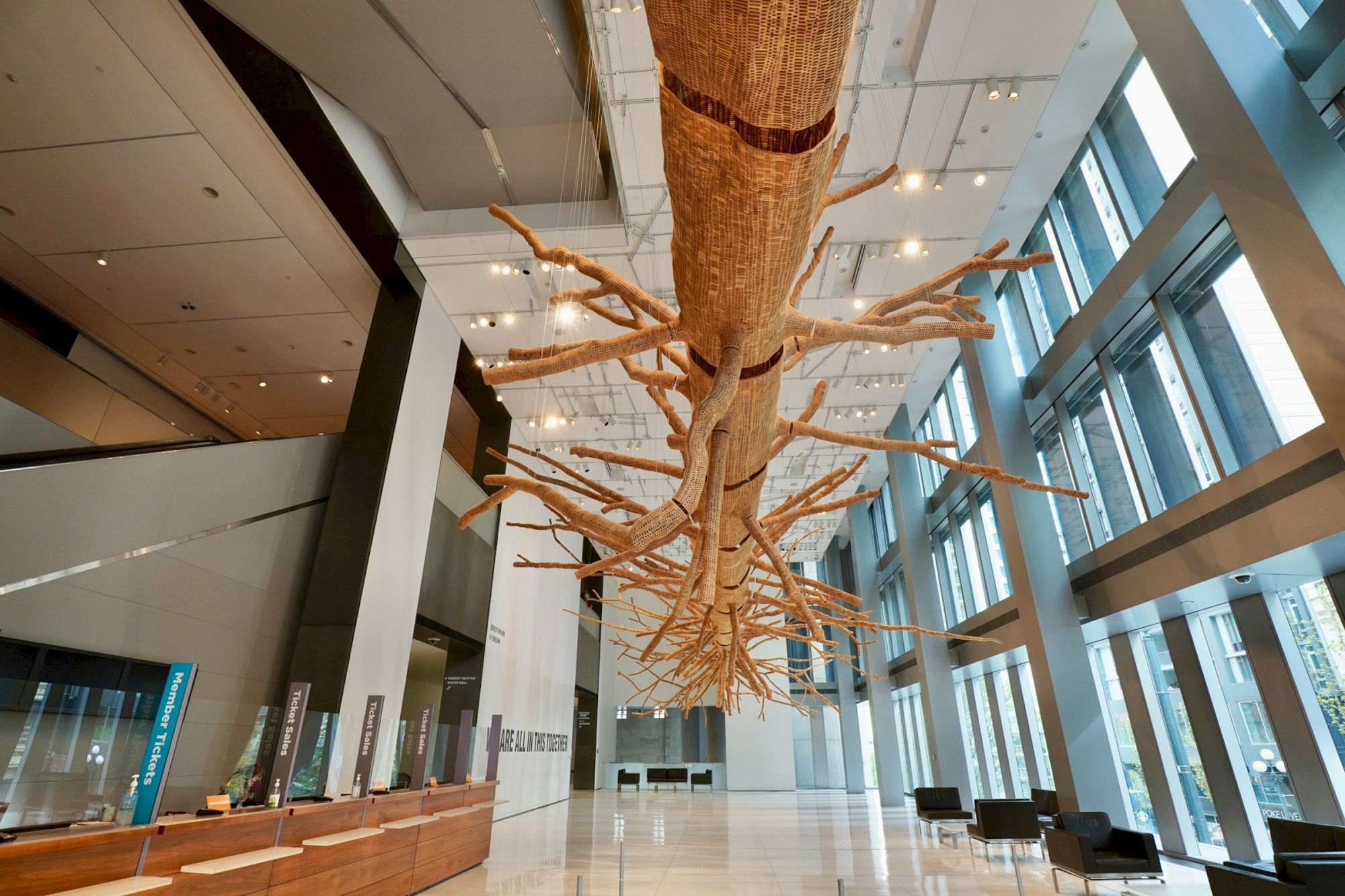 A large, suspended tree sculpture with sprawling branches hangs from a bright, modern hotel/office lobby ceiling. The space has tall windows and sleek seating areas, ending with a calm, airy vibe.