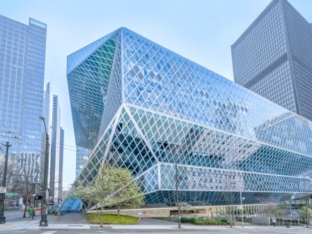 A futuristic glass building with a diamond-patterned fa&ccedil;ade sits among tall skyscrapers in a city, reflecting blue skies and street activity.