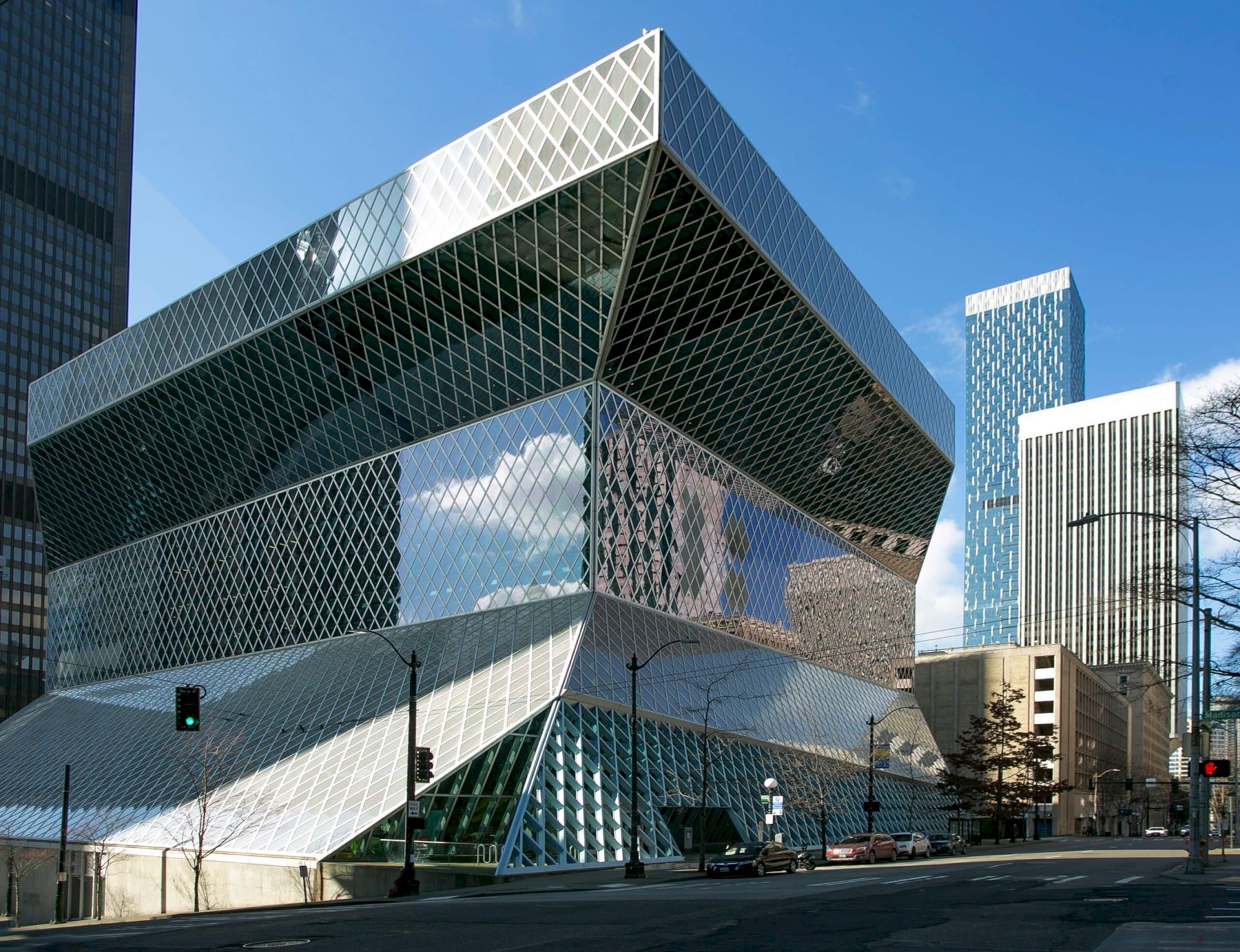 A futuristic, glass-and-metal building with stacked, angular blocks reflecting the blue sky, in a city street scene.