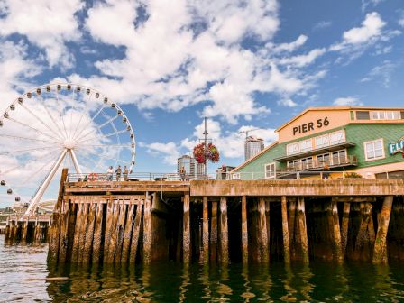 A seaside pier with weathered pilings, a large Ferris wheel, a green pier building labeled &ldquo;PIER 56,&rdquo; and a blue sky with scattered clouds.