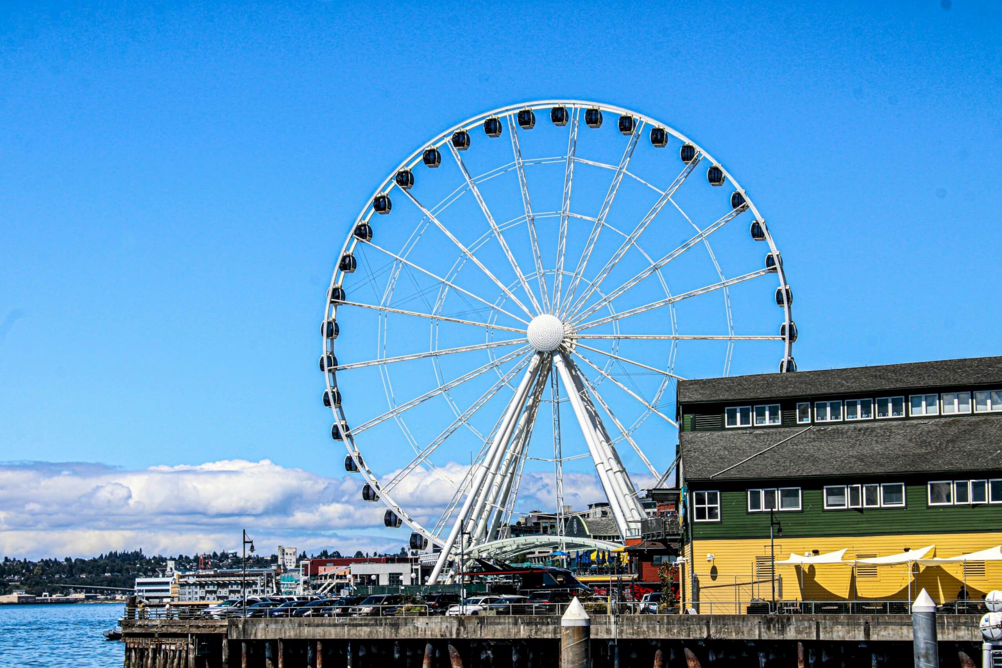 A large Ferris wheel on a pier beside a colorful building by the water, with a bright blue sky and calm sea in the background.