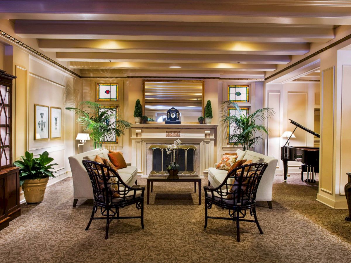 A cozy hotel lobby with a central fireplace, two white sofas facing each other, dark wooden chairs, plants, and warm ambient lighting.