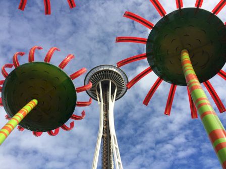 A view from below of colorful skyward structures near a tall tower, with bright red tentacle-like sculptures framing the blue sky.