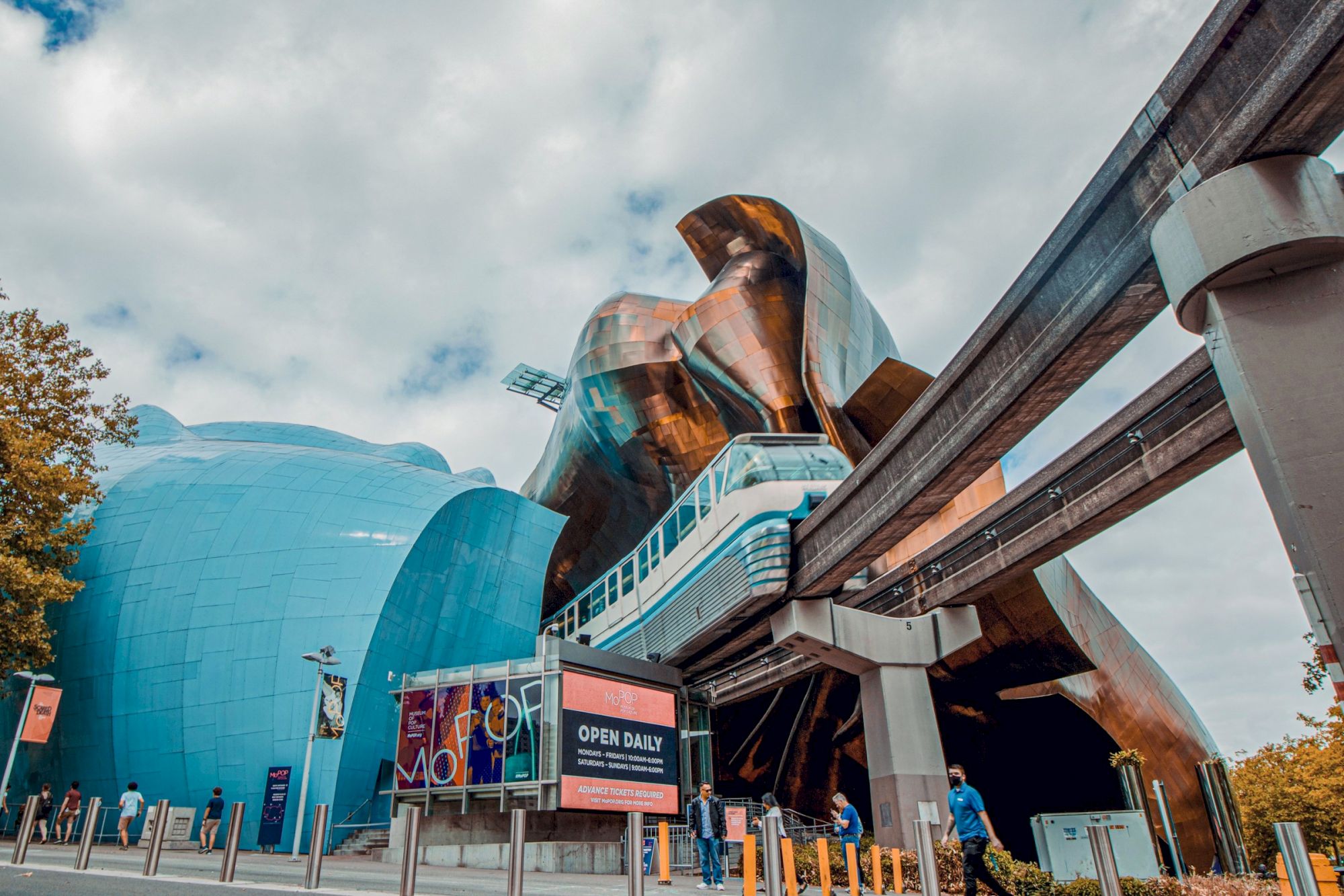 A futuristic urban scene with orange sculptures, a blue dome, monorail tracks, banners, and people near a ticket booth in a modern plaza.