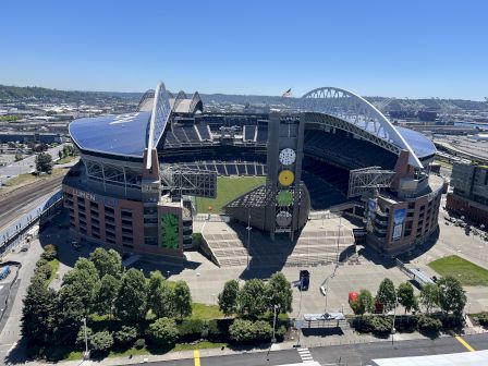Aerial view of a modern stadium with a curved, sail-like roof, green field inside, surrounding parking, trees, and a clear blue sky.