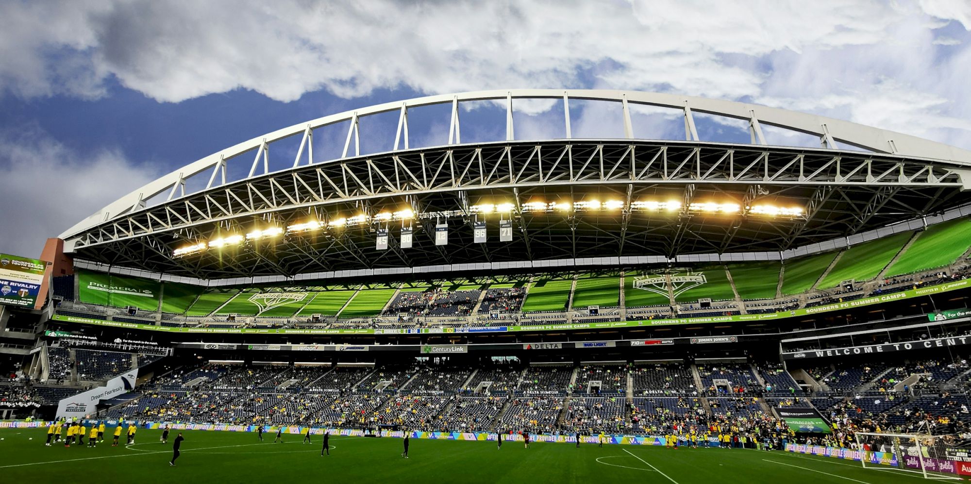 A large, modern stadium with a curved white roof, bright floodlights, green seats, and a soccer field filled with players and spectators, under a blue sky.