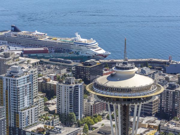 A cityscape with high-rise buildings and a large space-ship-like tower, plus a cruise ship docked at a harbor in the background, overlooking blue water.