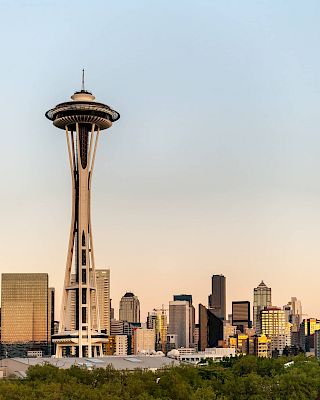 A skyline view of Seattle featuring the iconic Space Needle rising above modern buildings at sunset.
