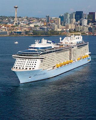 A large white cruise ship sails near a city skyline, with a tall tower and high-rise buildings in the background, on calm blue water.