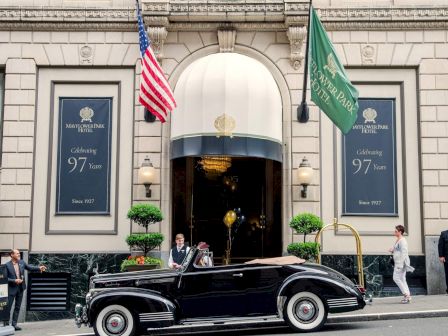 A vintage black car pulls up at a grand hotel entrance, flags flying, with uniformed staff and guests in front. There are blue banners and potted trees flanking the doorway.