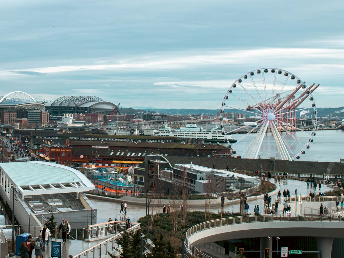 A bustling waterfront with a Ferris wheel, boardwalk, shops, and a large observation deck by the water.