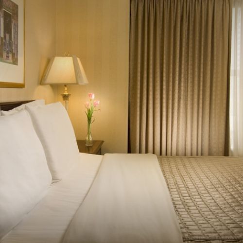 A hotel room with a neatly made double bed, white pillows, bedside lamp, framed art, and a window with drapes&mdash;calm and cozy ambience.