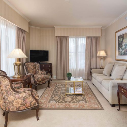 A luxurious living room with beige walls, cream sofa, floral armchairs, a coffee table, lamps, and a framed artwork; curtains and a flat-screen TV complete the setup.