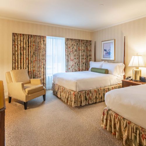 A hotel room with two large beds, a seating area, dresser with TV, nightstands, lamps, patterned curtains, and soft beige decor.