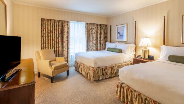 A hotel room with two double beds, a flat-screen TV, an armchair, and a window with curtains.