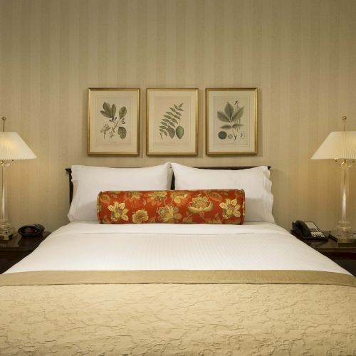A neatly made hotel bed with white linens, a decorative red floral bolster, two nightstands, lamps, and framed botanical prints above the headboard.