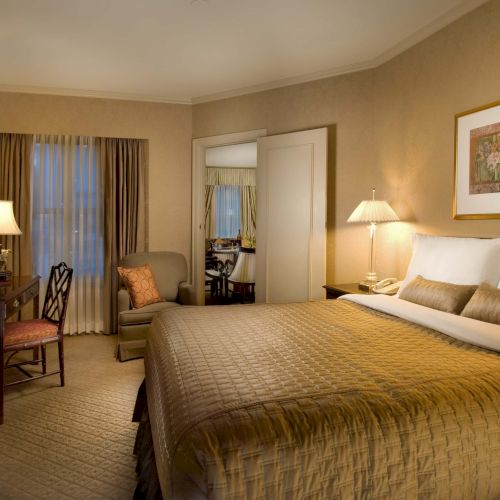 A hotel bedroom with a large bed, two nightstands, desk and chair, a TV dresser, warm lighting, and framed artwork on the wall.