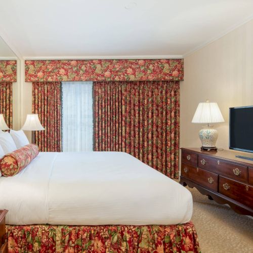 A tidy hotel room featuring a large bed with white linens, floral curtains, matching bed skirt, and a dresser with a flat-screen TV. ending.
