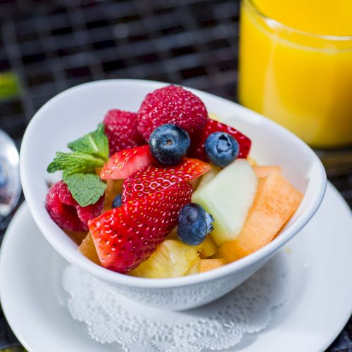 Fresh fruit bowl with strawberries, blueberries, melon, pineapple, and mint, plus orange juice in the background.